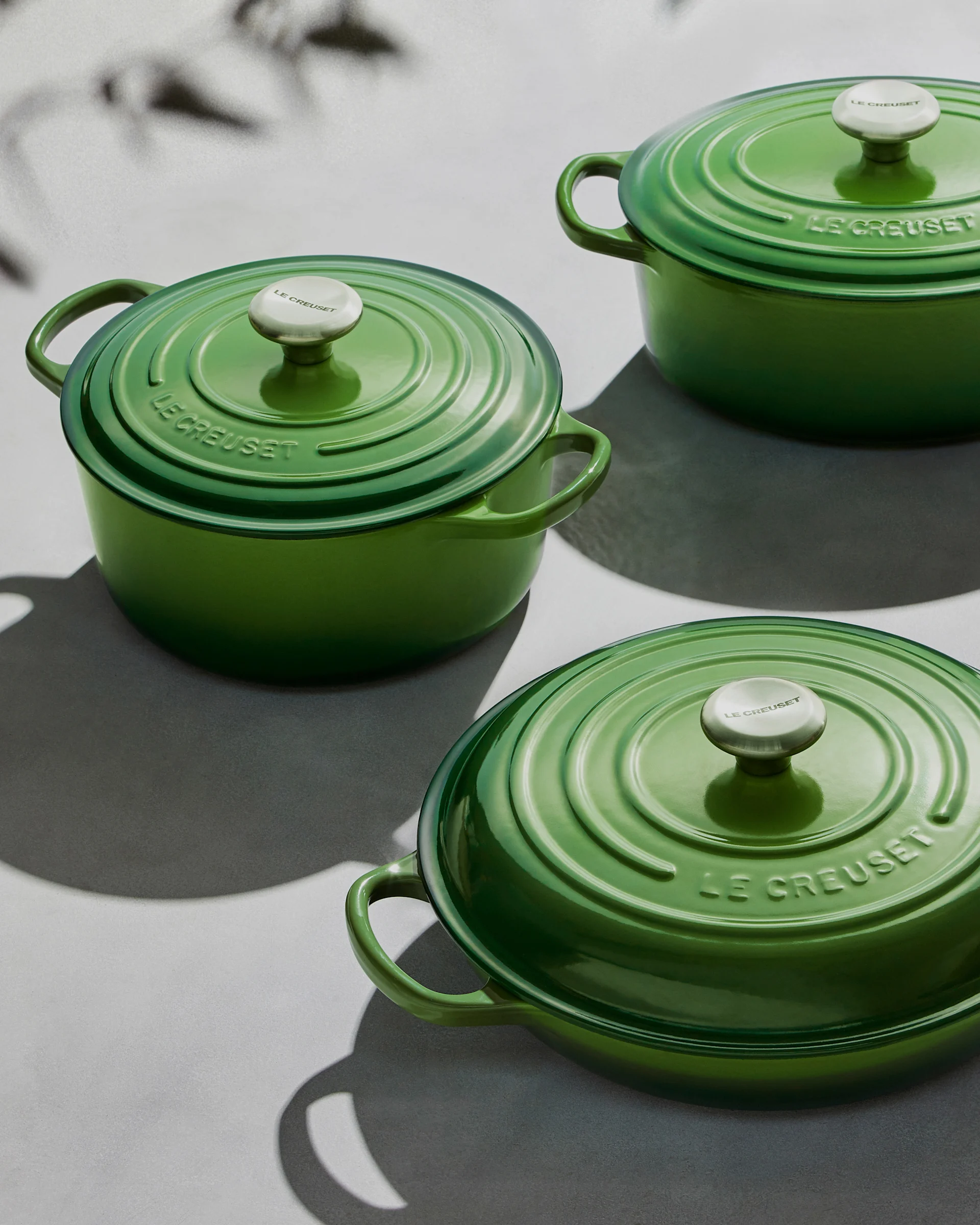 Three green Le Creuset pots with lids and silver knobs on a light surface with shadows.