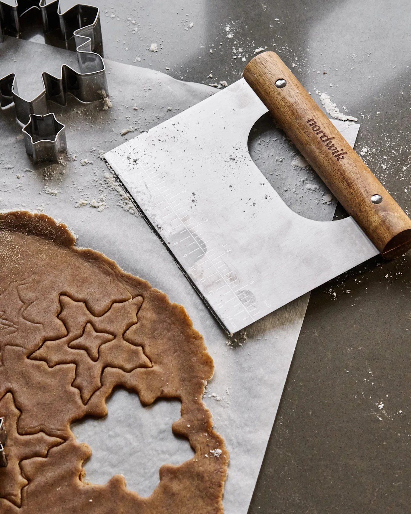 Gingerbread dough with star and leaf cutouts, metal cookie cutters, and a dough scraper on a flour-dusted surface.