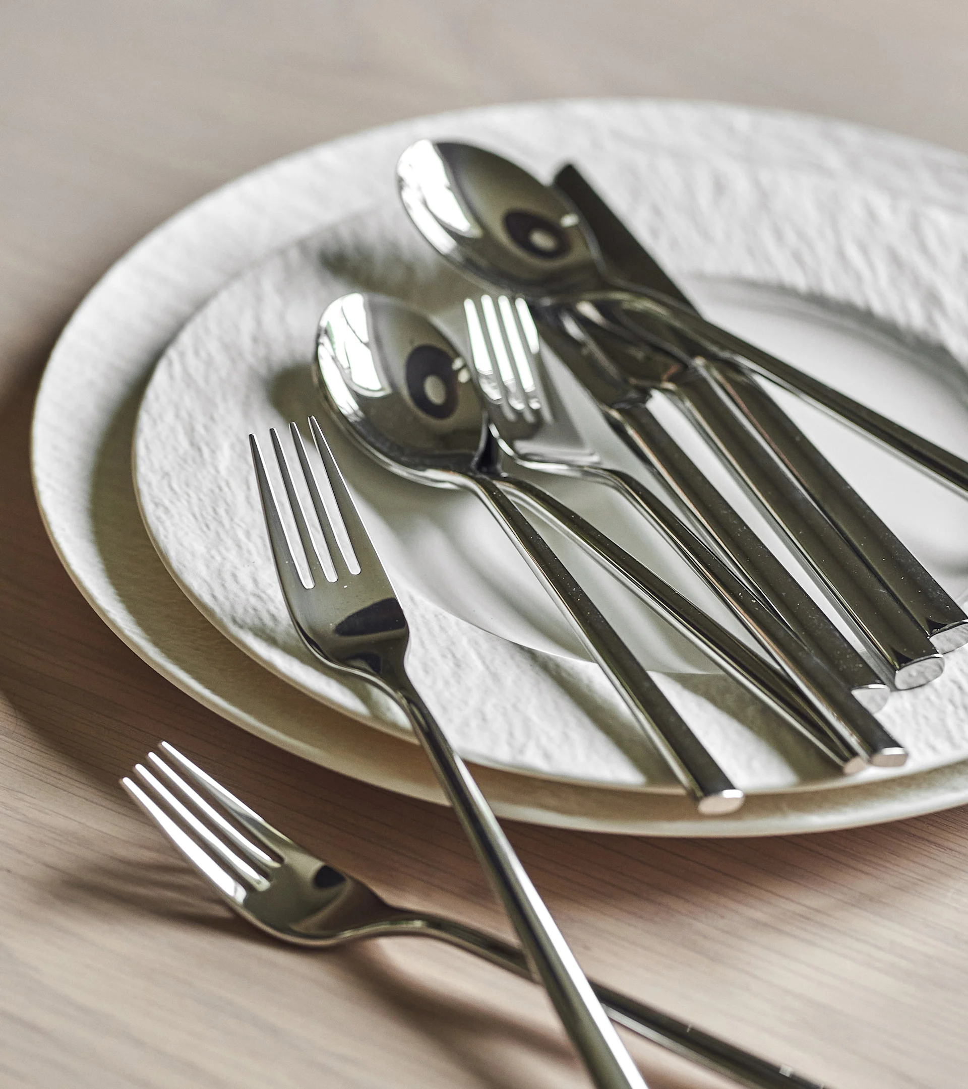 Shiny silver cutlery set on two white textured plates on a light wooden table.
