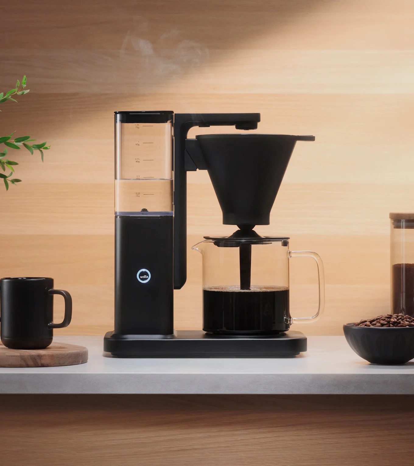 A black coffee maker brewing coffee into a glass carafe on a light counter, with steam rising. A black mug and coffee beans are nearby.