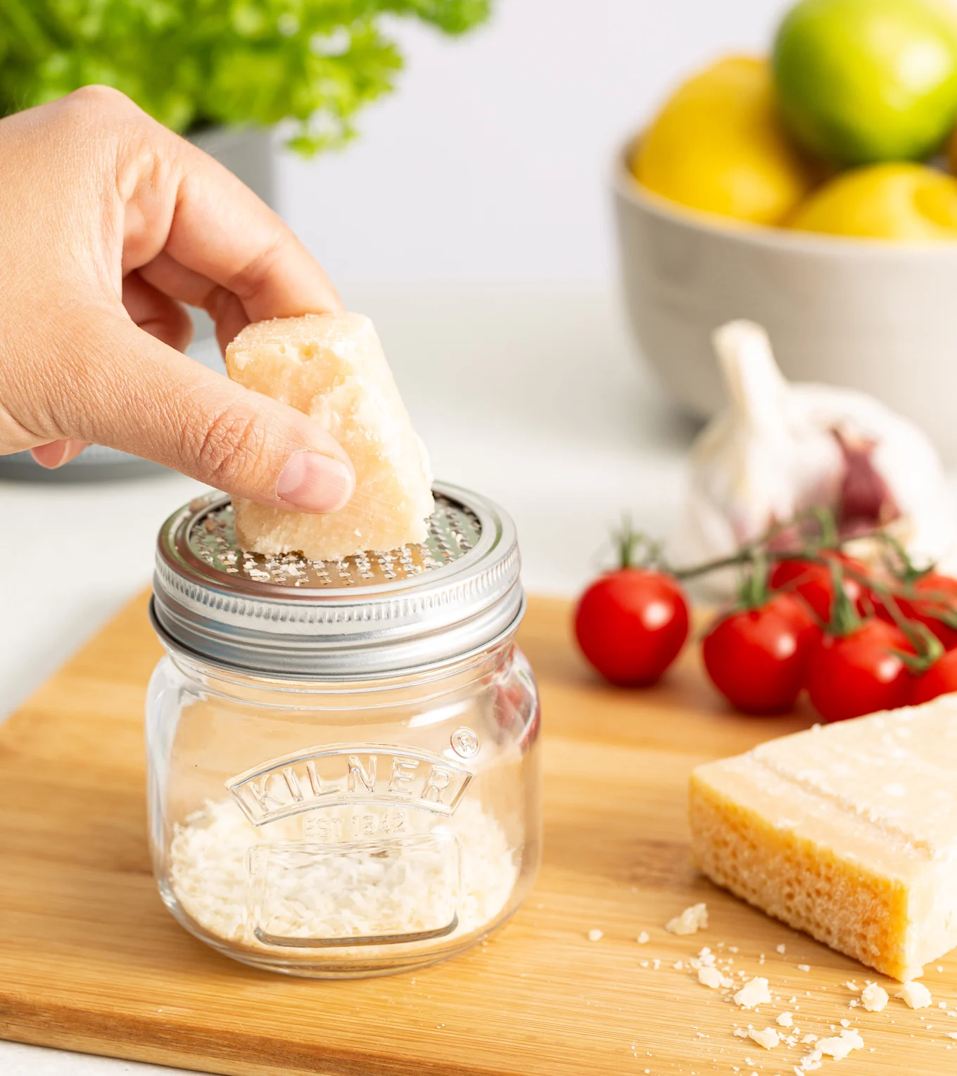Hand grating hard cheese into a clear glass jar with a grater lid. Fresh tomatoes, garlic, and a cheese block are on a wooden board.
