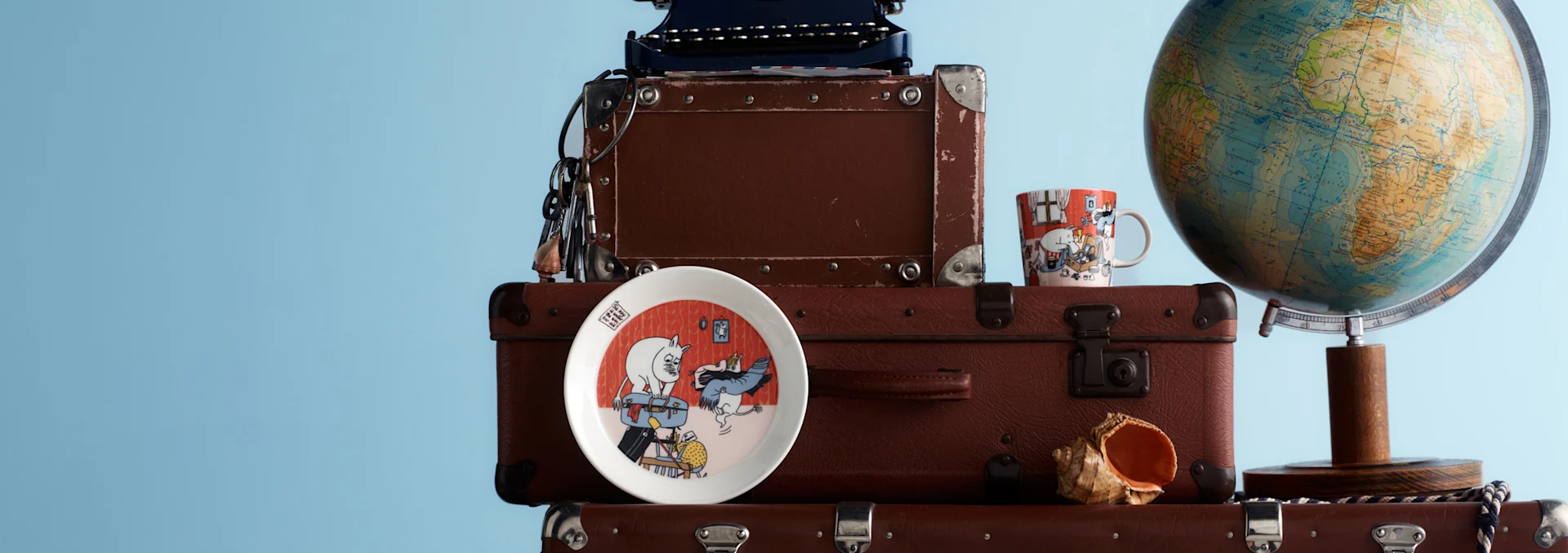Stacked brown vintage suitcases with a blue typewriter, Moomin plate and mug, seashell, and a globe, against a light blue background.