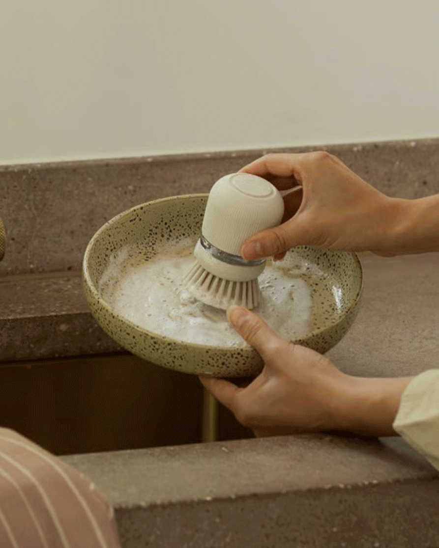 Hands washing a speckled ceramic bowl with a white dish brush in a sink.