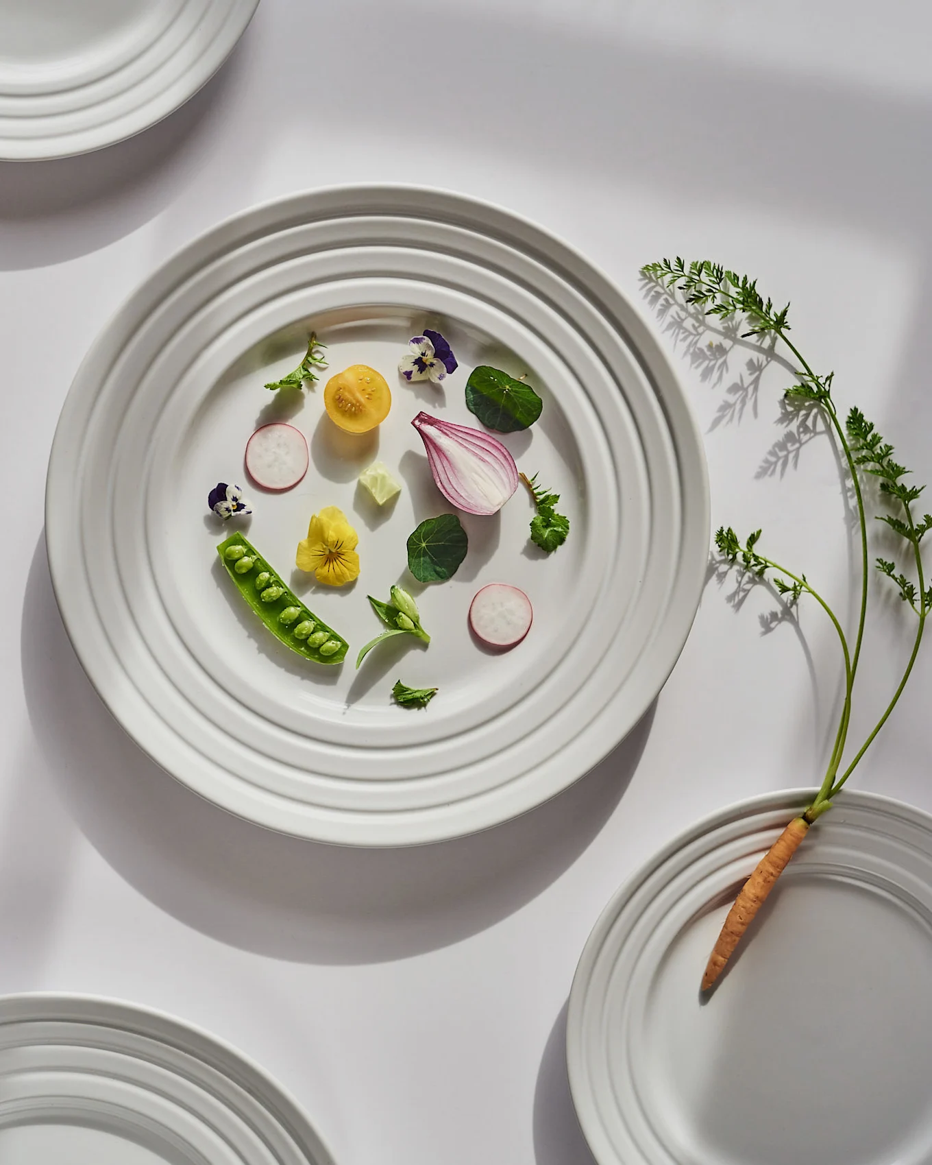 A white plate displays fresh cut vegetables and edible flowers, with a whole carrot and its greens on another plate.