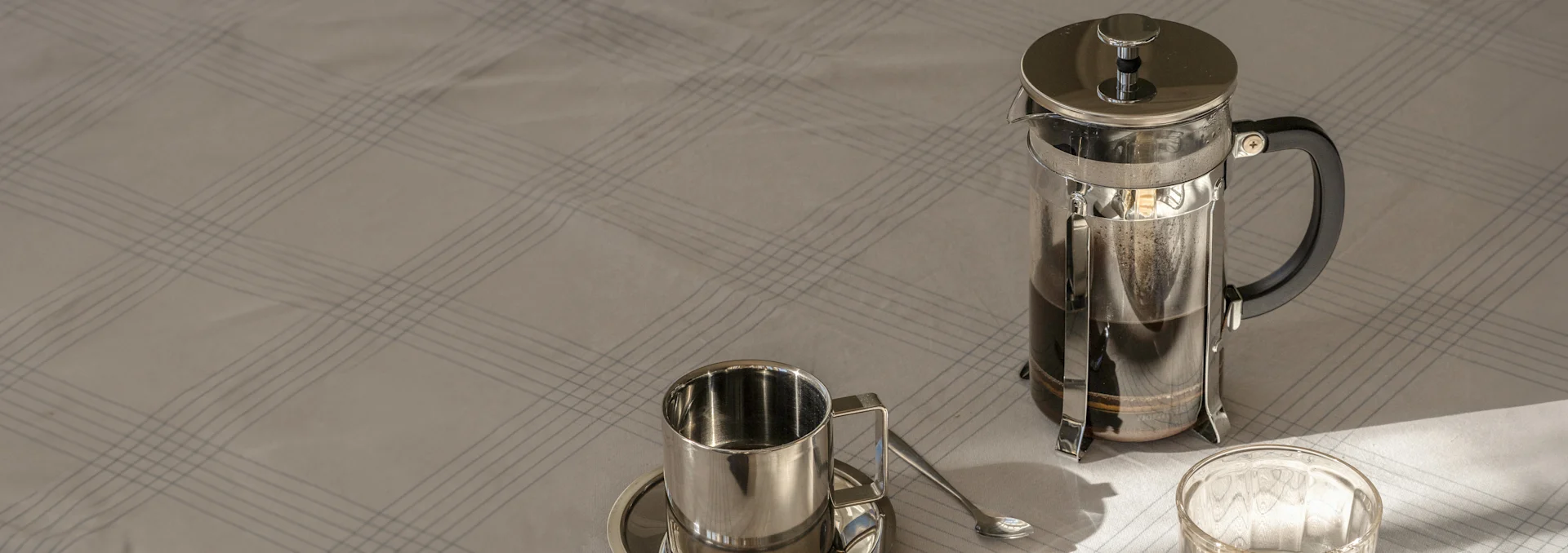 French press with coffee, a metal cup and saucer with a spoon on a light, plaid tablecloth in sunlight.