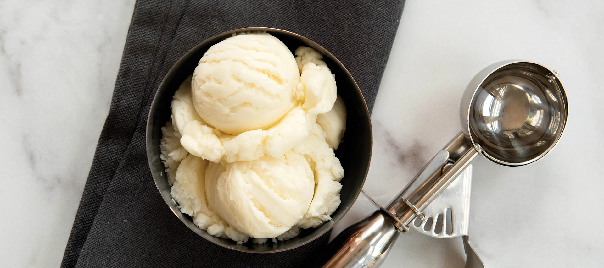 Scoops of vanilla ice cream in a dark bowl next to an ice cream scoop on a marble surface.