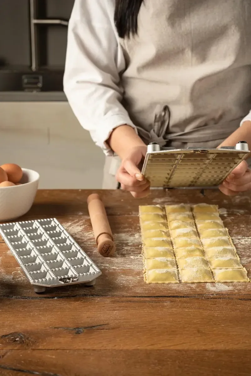 Person making ravioli with a metal ravioli maker, fresh ravioli, a rolling pin, and eggs on a wooden table.