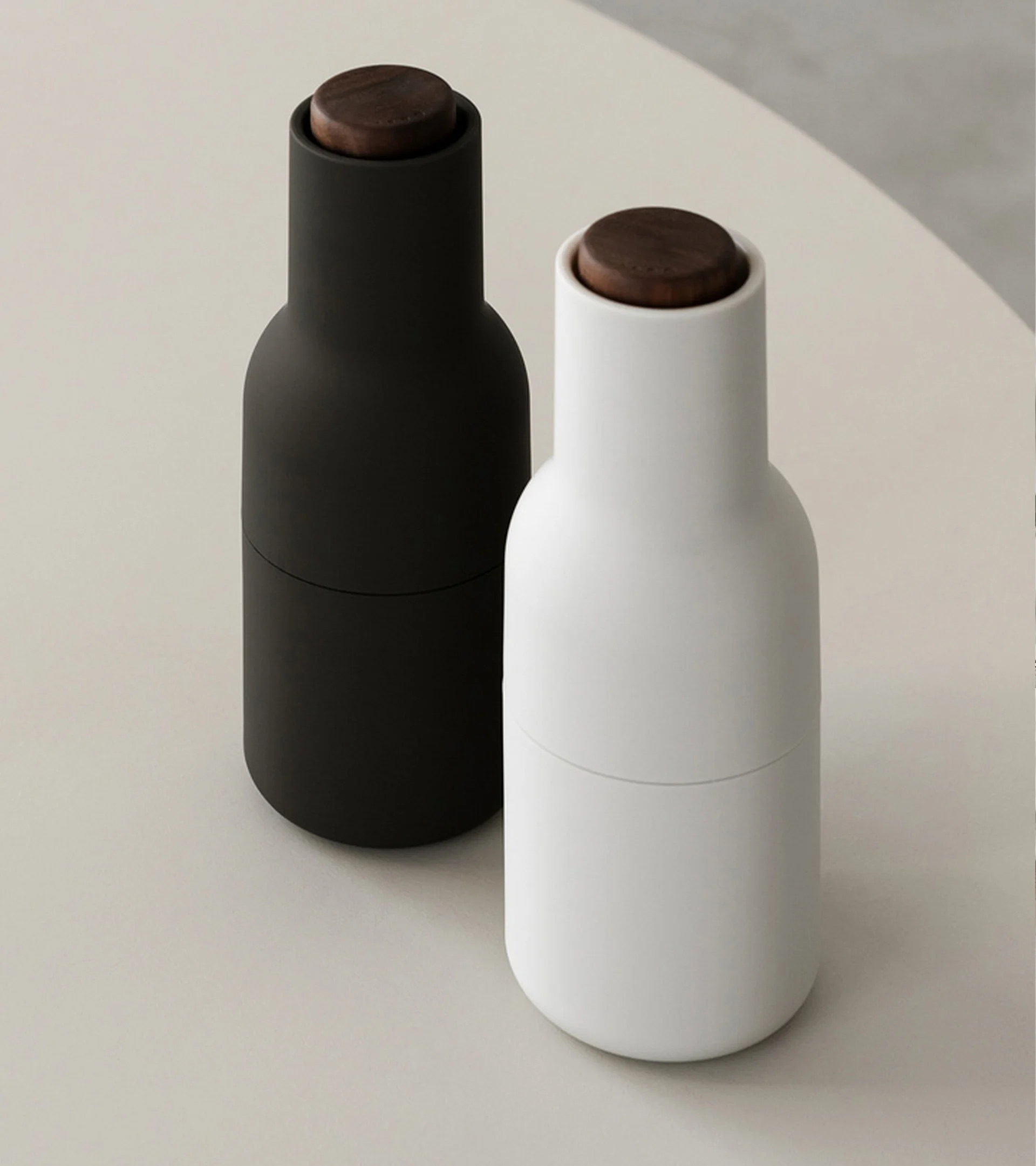 Two minimalist cylindrical grinders, one black and one white, with dark wooden tops.