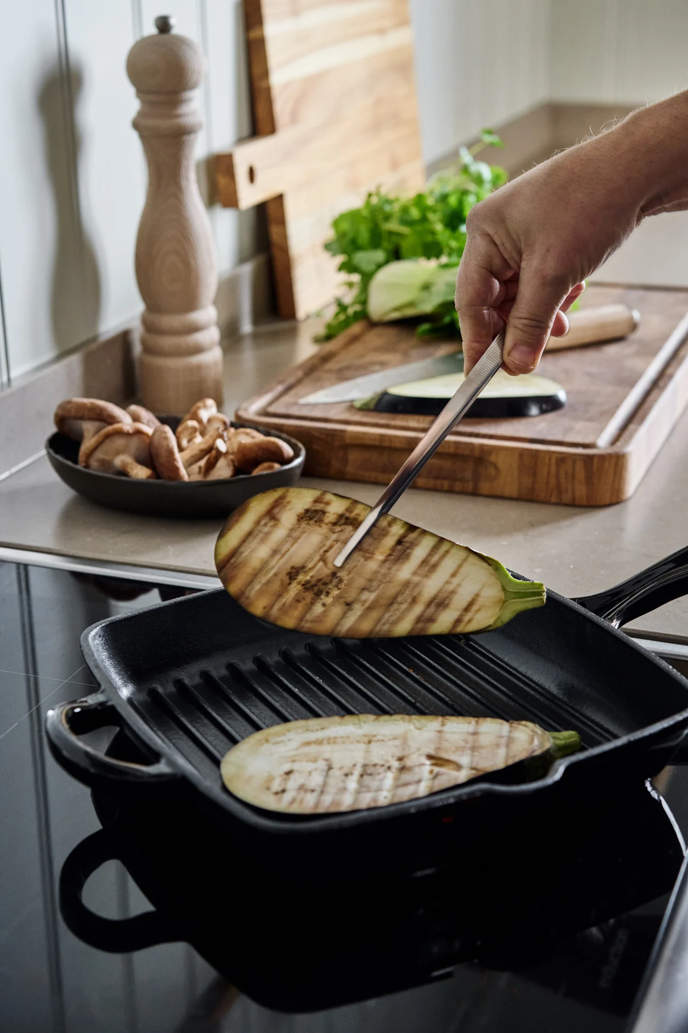 Hand flipping grilled eggplant slices in a black cast iron grill pan on a modern stovetop. Ingredients are in the background.