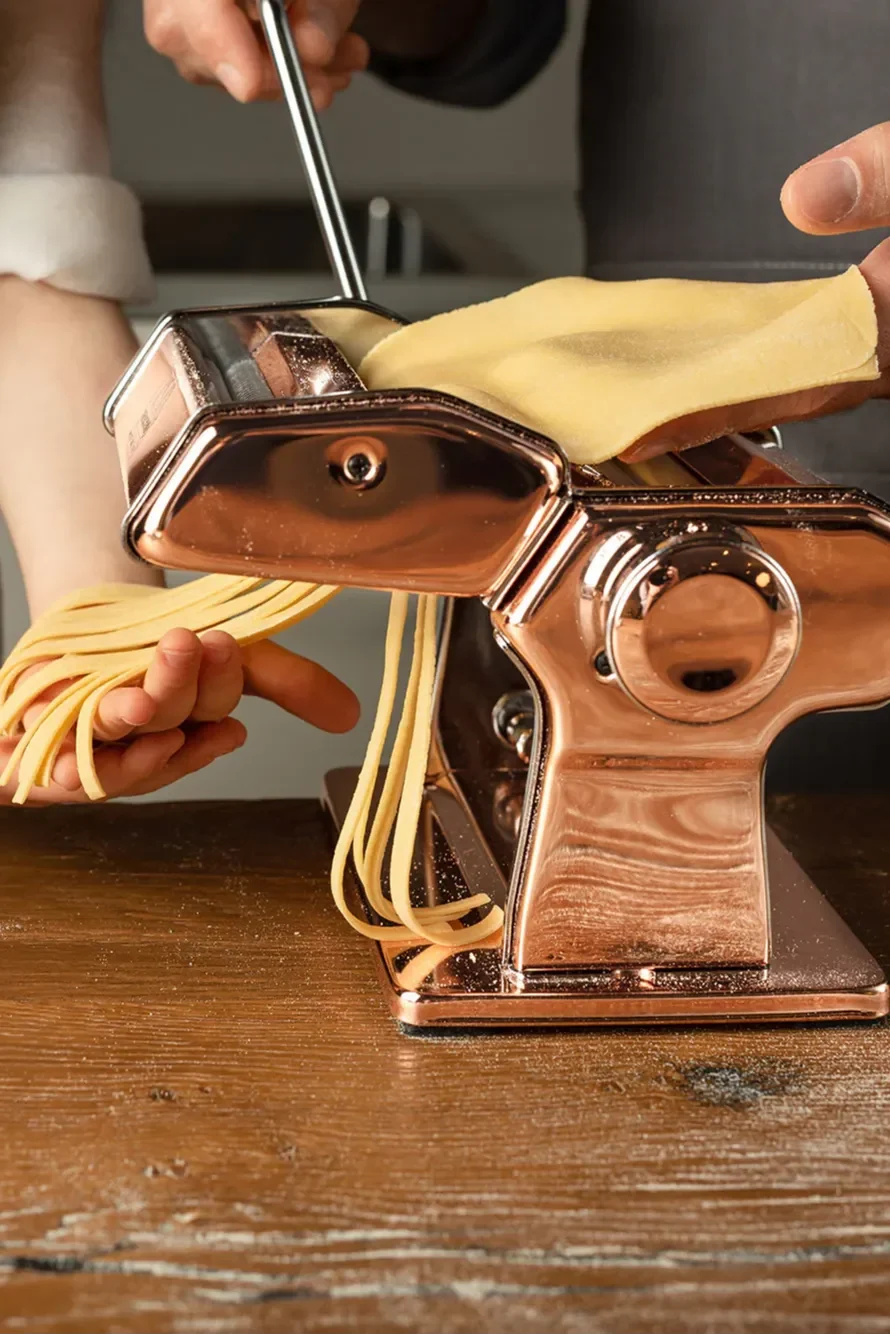 Hands operate a copper pasta machine, feeding dough and producing fresh pasta strands on a wooden surface.