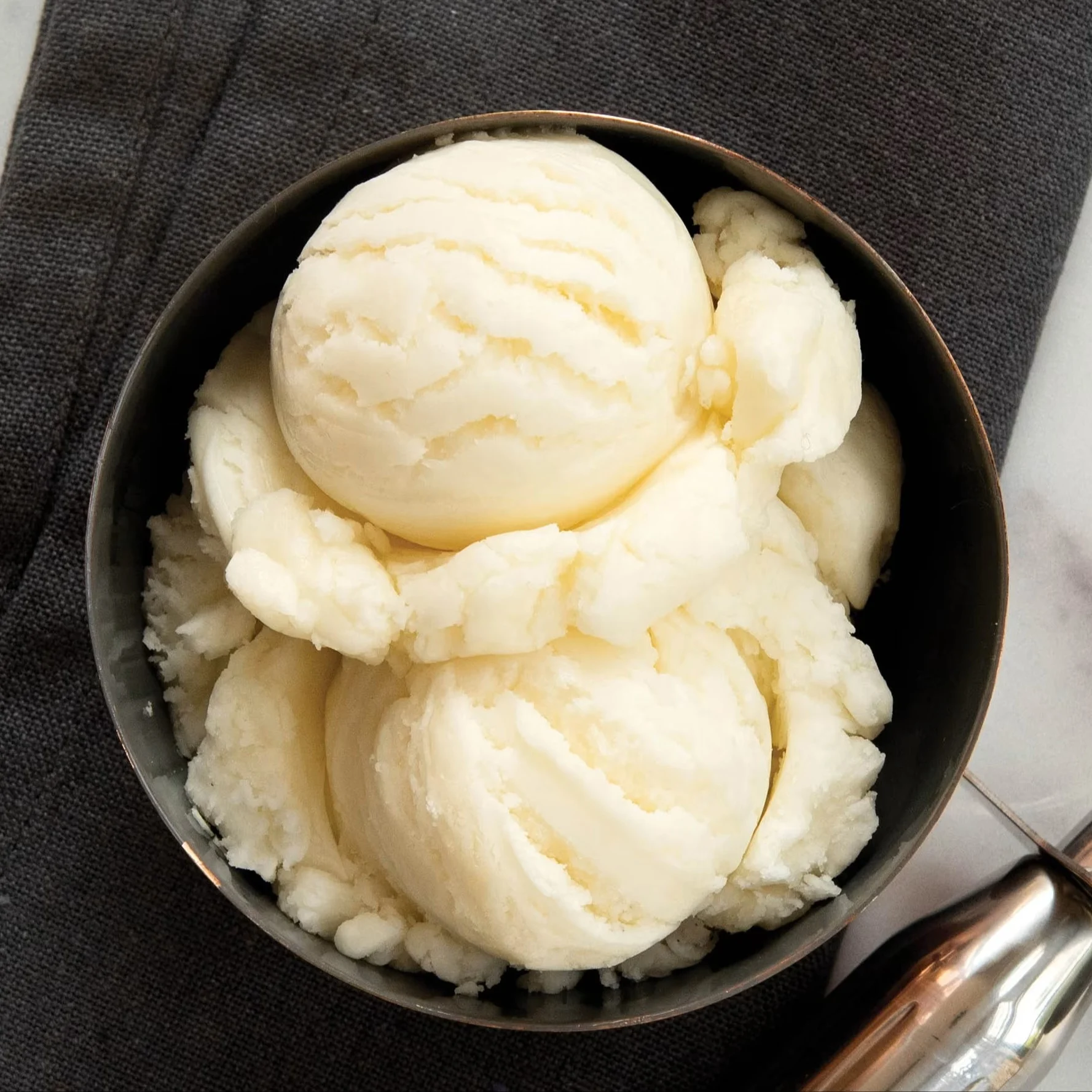 Scoops of light-colored ice cream in a dark bowl on a dark gray cloth, with an ice cream scoop nearby.