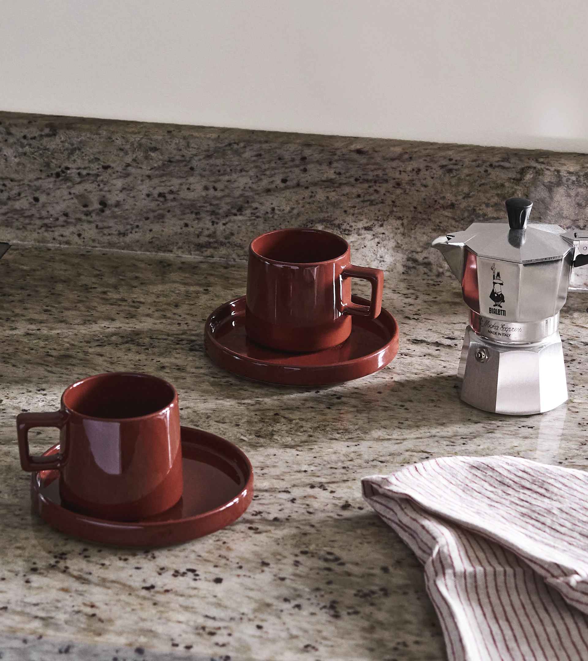 Two red-brown coffee cups with saucers and a silver Moka pot on a speckled countertop, with a striped cloth.