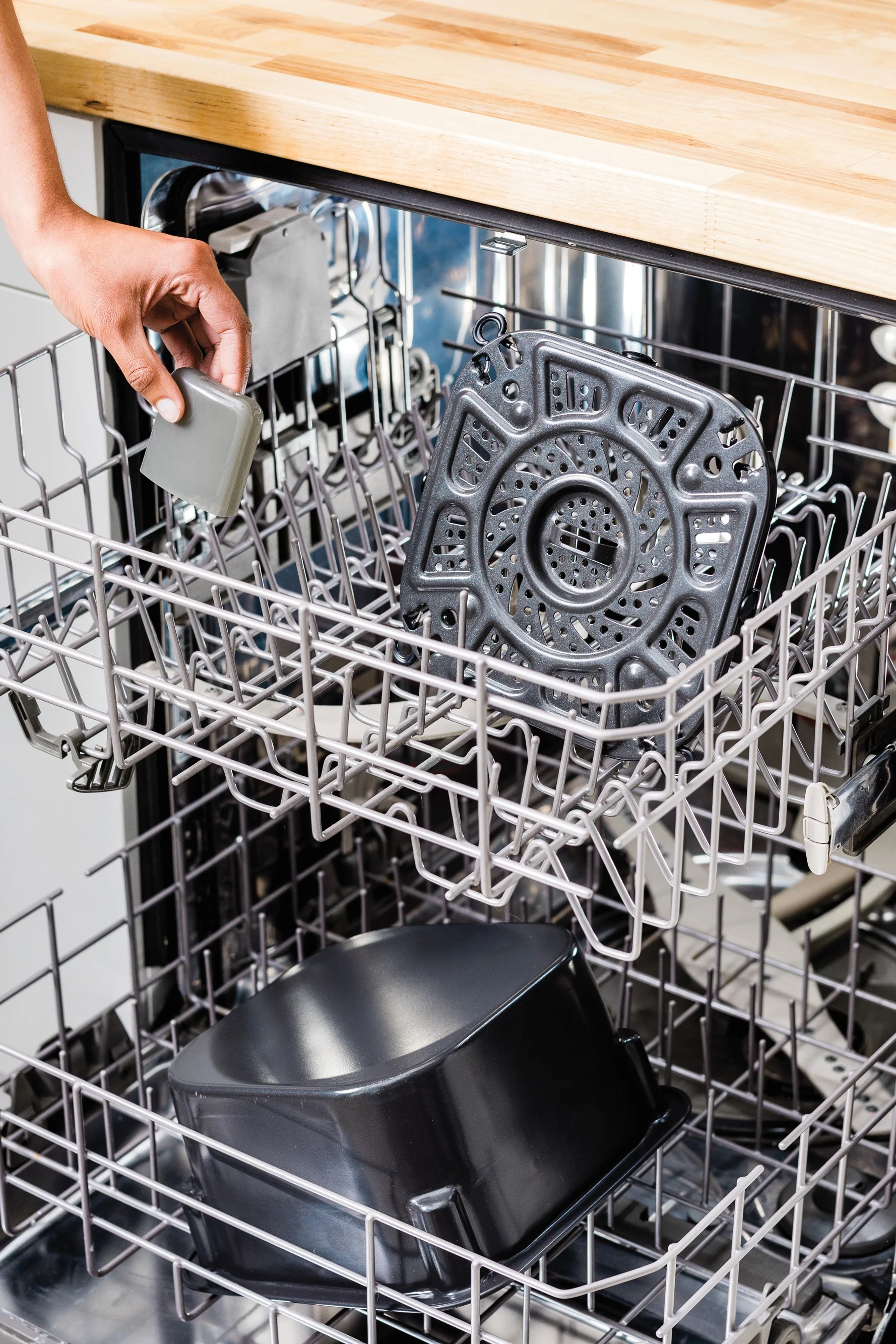 A hand places a small gray item into an open dishwasher, which holds a dark gray air fryer basket and pot.