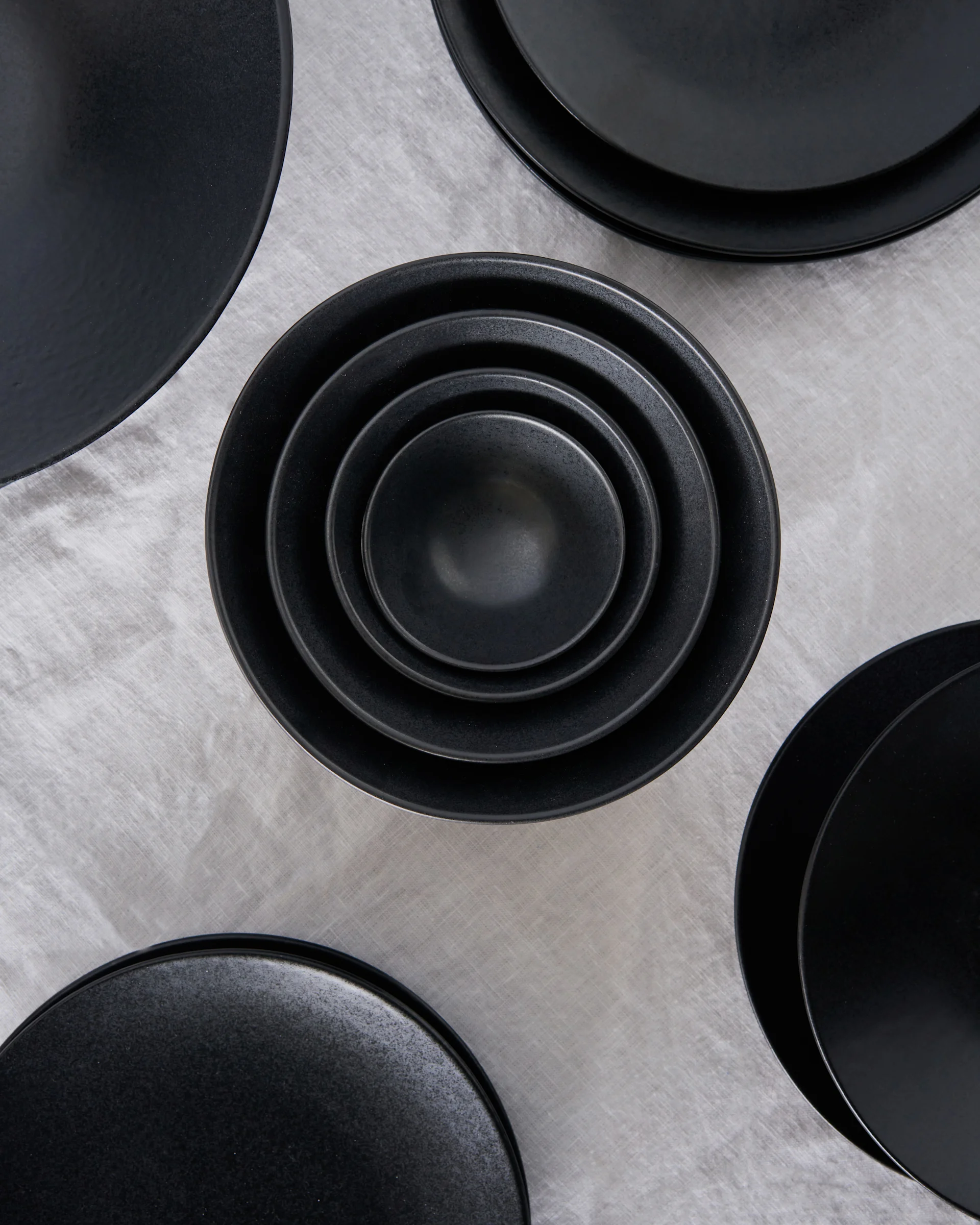 Overhead shot of a set of black ceramic bowls stacked concentrically in the center, surrounded by other black plates on a grey linen tablecloth.
