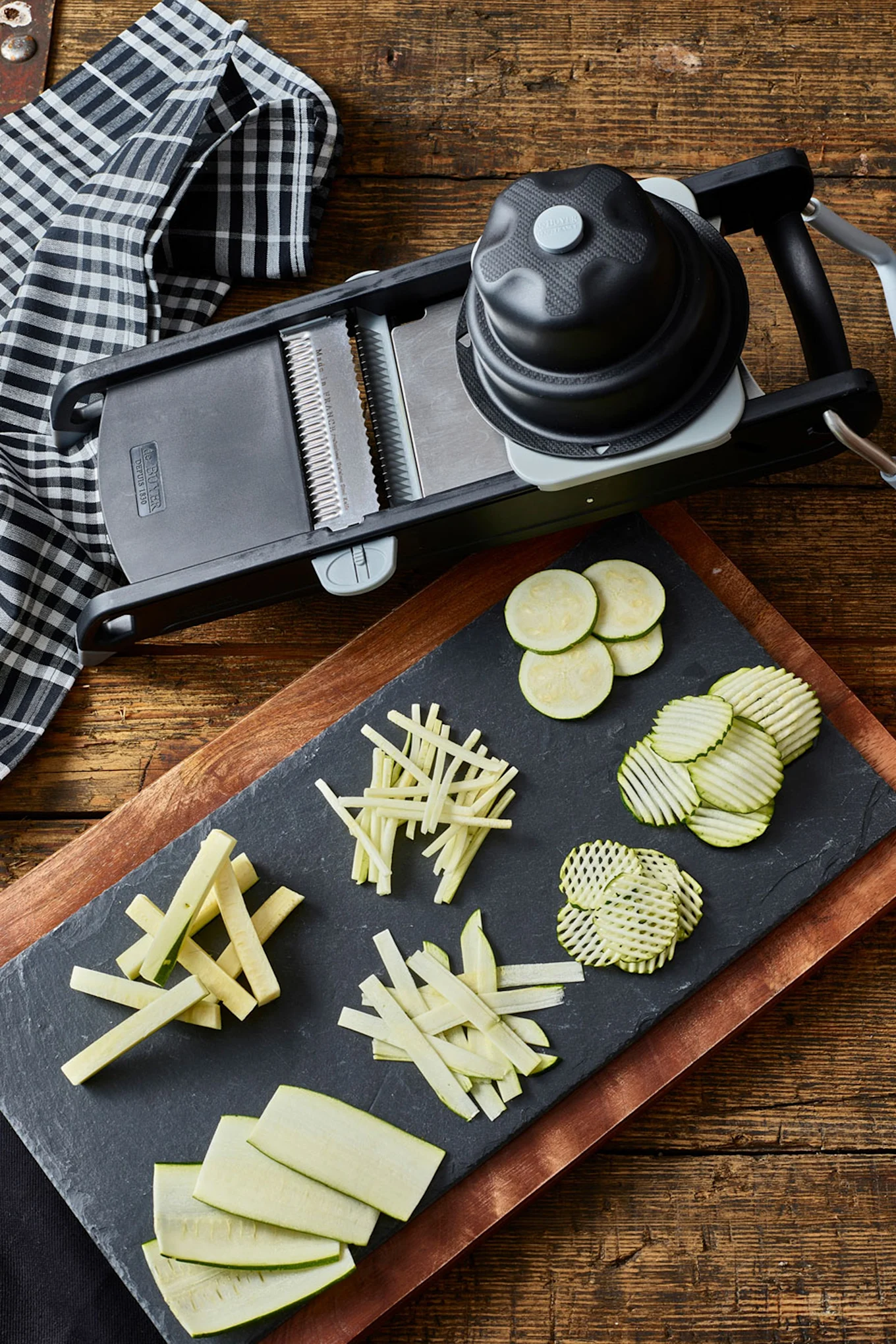 A hand operates a mandoline slicer, neatly collecting sliced cucumbers in a pull-out drawer on a wooden board.