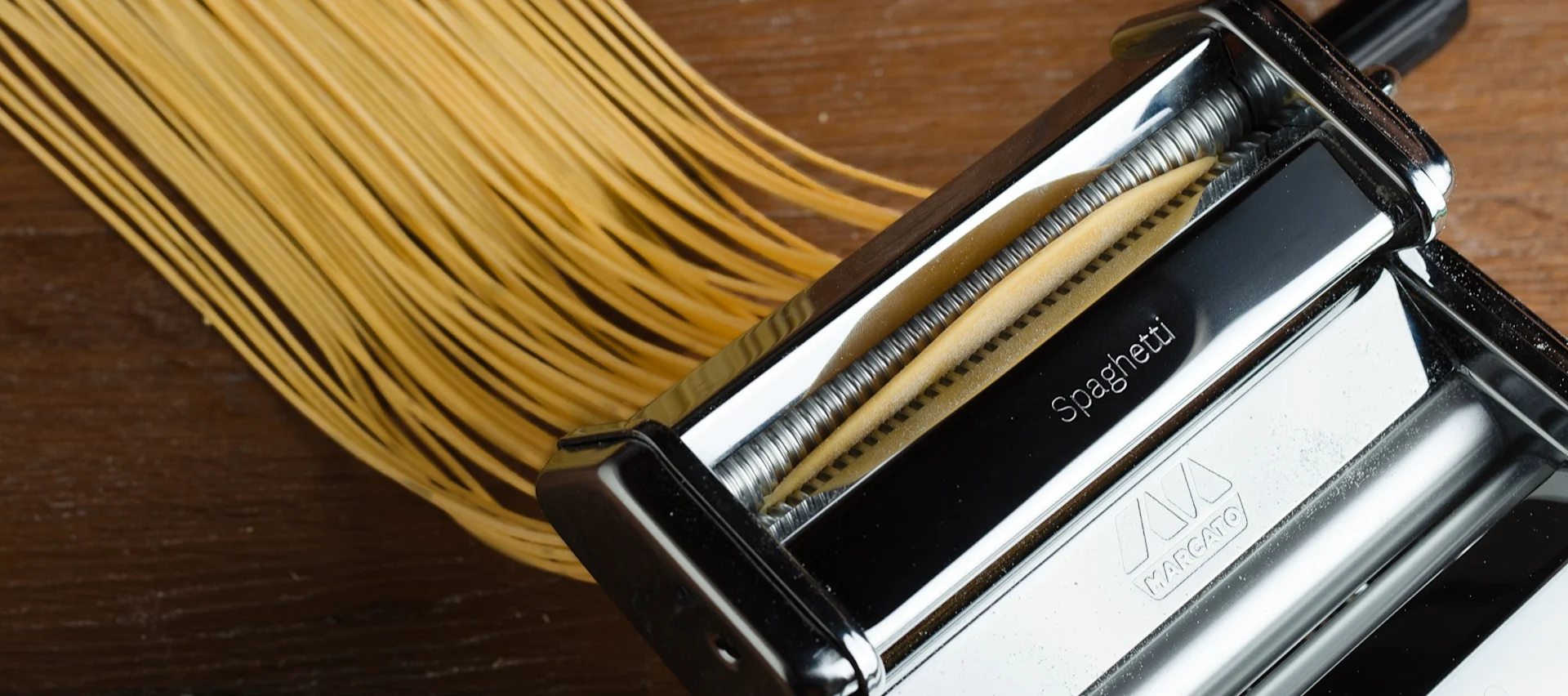 Fresh spaghetti being extruded from a pasta maker machine onto a wooden surface.