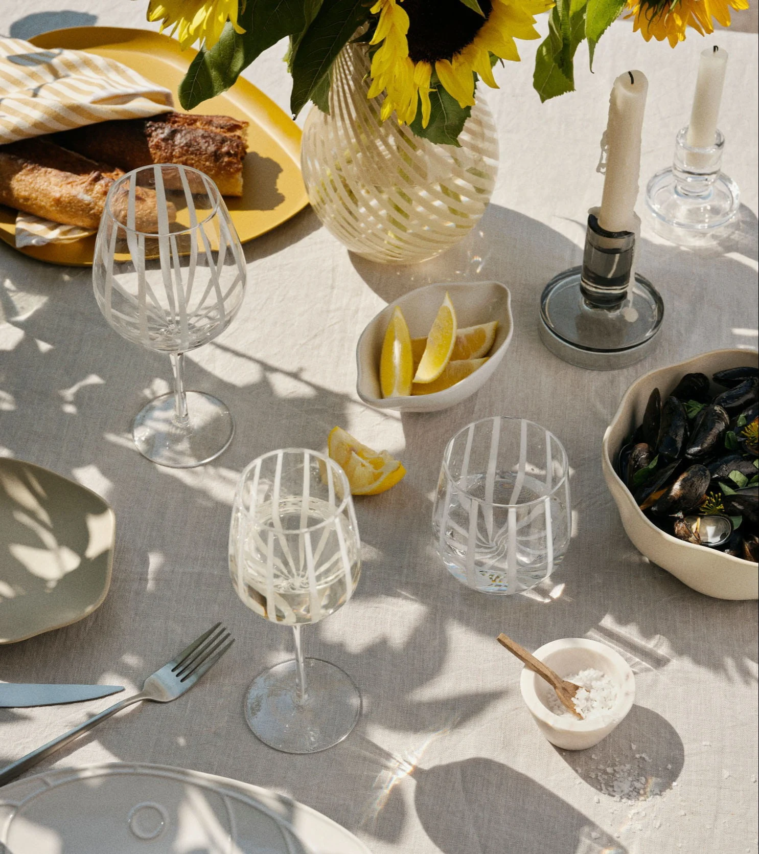 A sunny outdoor table setting with baguettes, mussels, lemons, wine glasses, sunflowers, and candles.