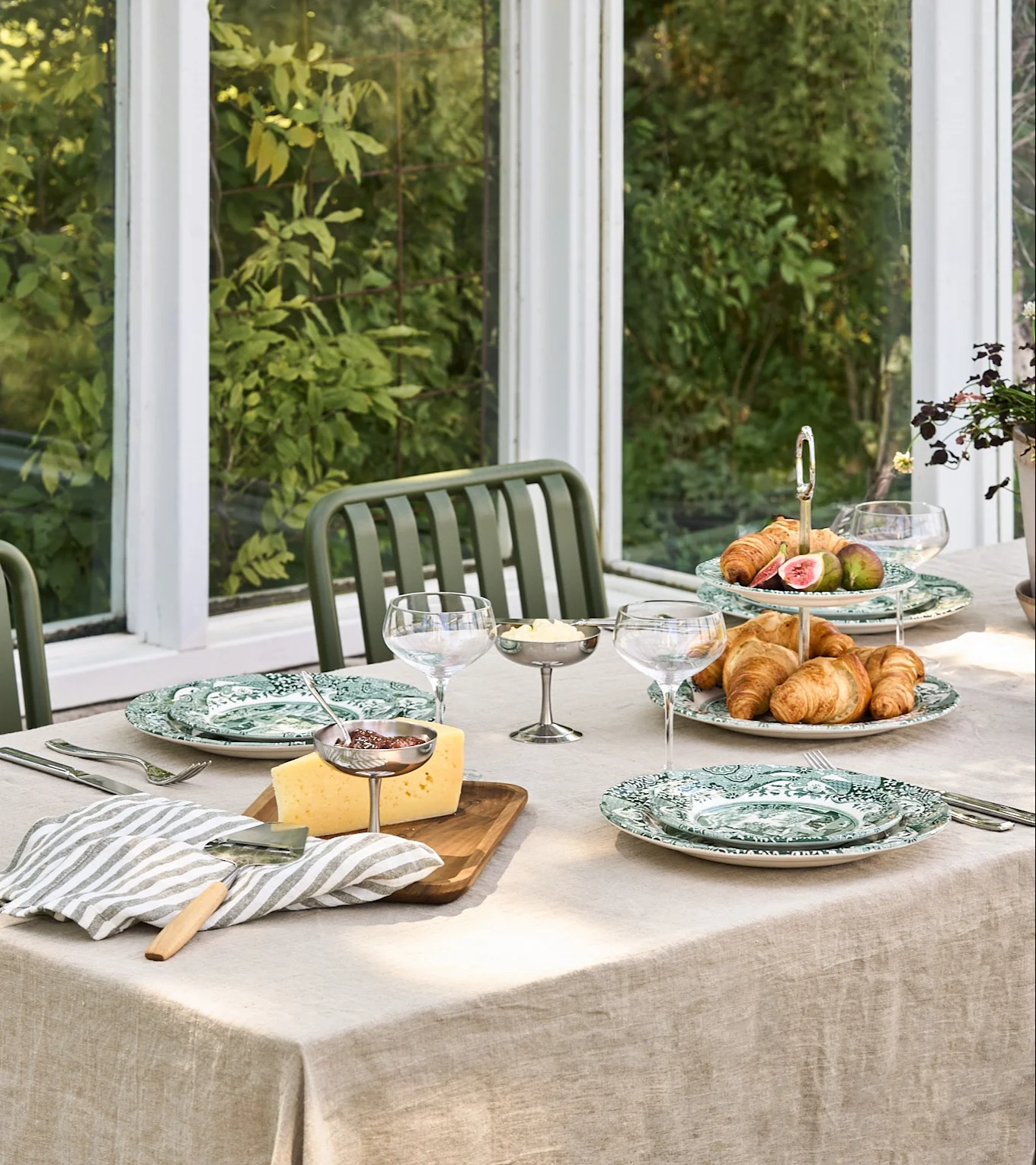 A breakfast table set with green patterned plates, croissants, figs, cheese, jam, and butter in a bright conservatory.