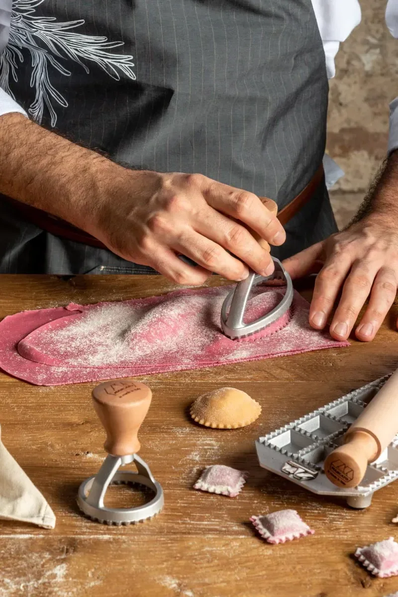 Hands using a pasta cutter on pink dough dusted with flour, making ravioli on a wooden table with tools.