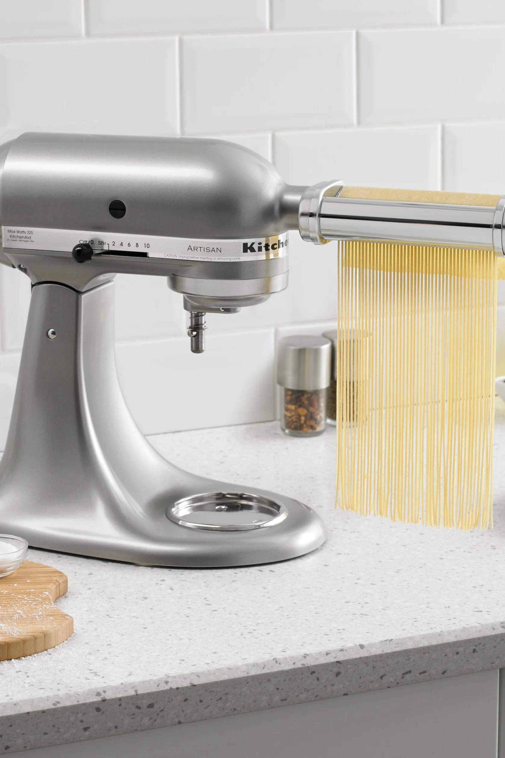 A silver stand mixer with a pasta maker attachment extruding long strands of fresh spaghetti onto a light gray speckled counter.