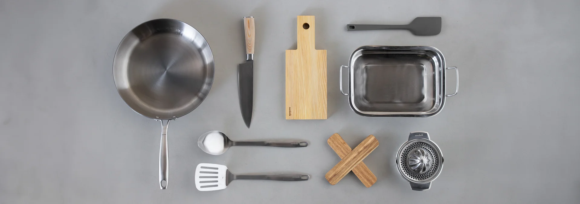 A flat lay of various kitchen utensils on a grey background: a frying pan, knife, cutting board, roasting pan, spatulas, ladle, wooden trivet, and citrus juicer.
