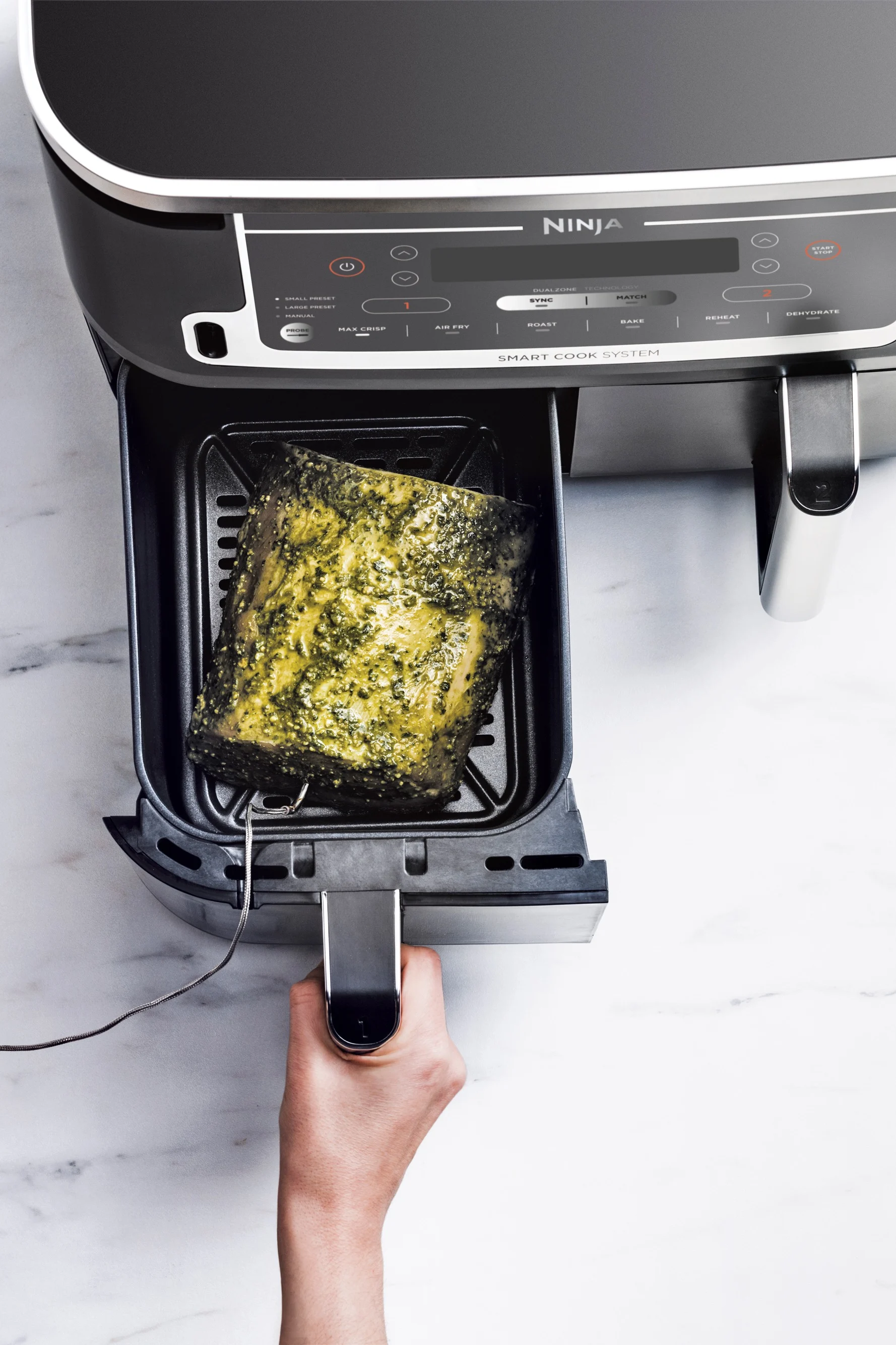 Hand pulling out an air fryer drawer with a herb-marinated roast and a meat probe inserted.