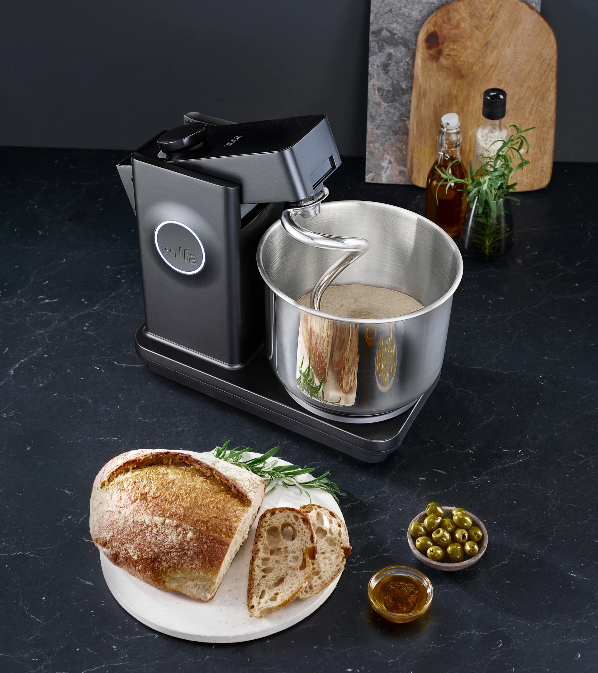 Black kitchen machine kneading dough in a stainless steel bowl, with a rustic bread loaf, sliced bread, olives, and olive oil on a dark counter.
