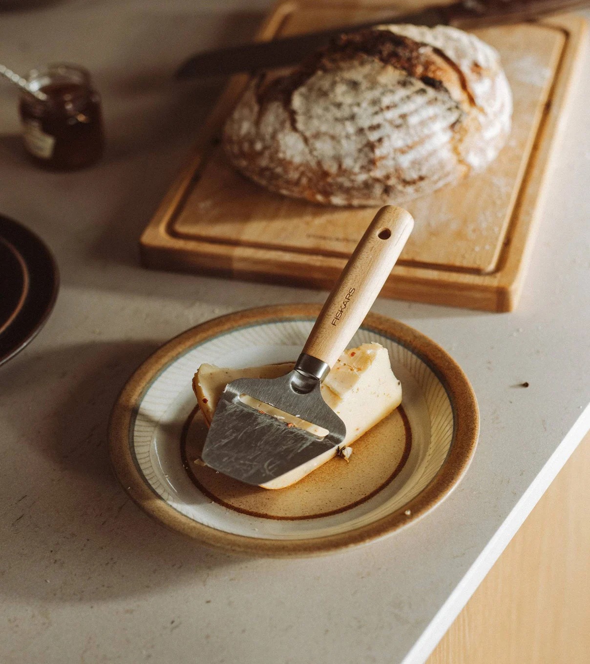 A cheese slicer with a wooden handle resting on a block of cheese on a plate, with a loaf of bread in the background.