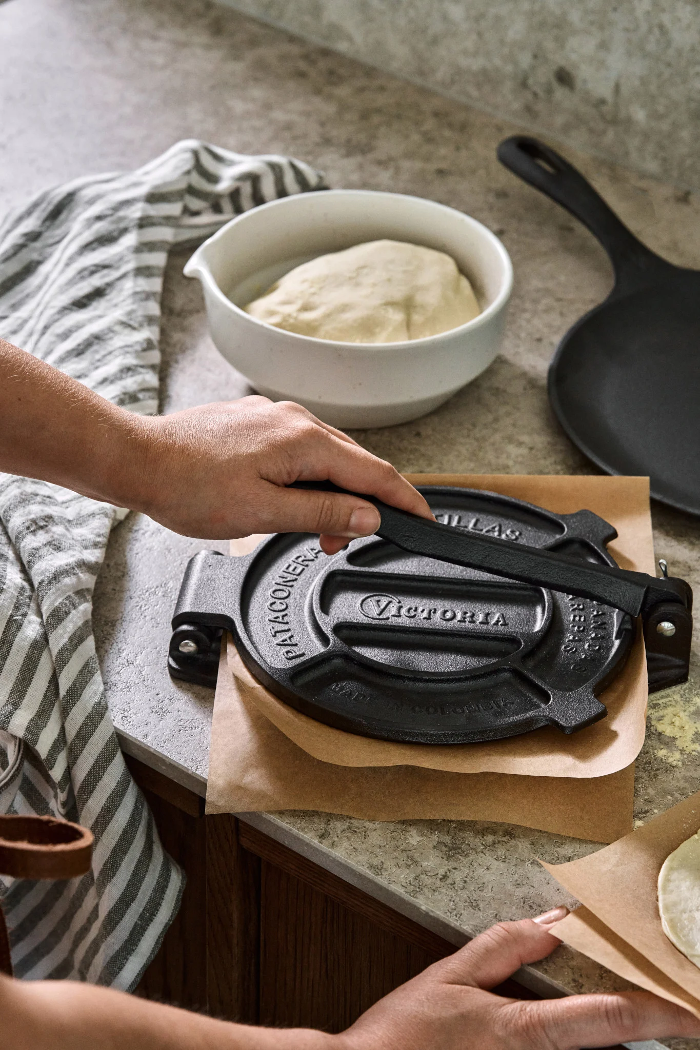 A hand presses dough in a cast iron tortilla press next to a bowl of dough and a striped tea towel. 