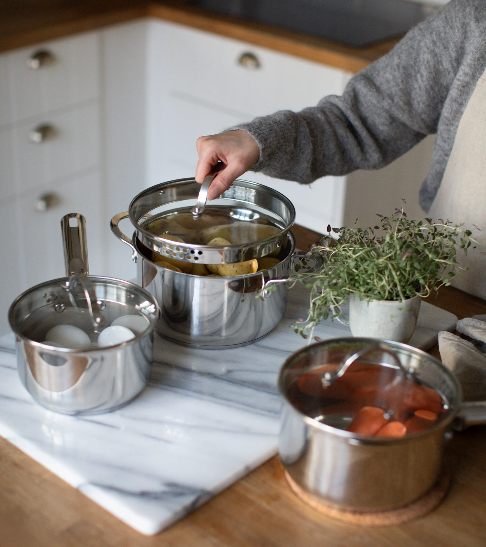 Hand lifting lid from a stainless steel steamer pot with potatoes, with eggs in another pot and carrots in a third.