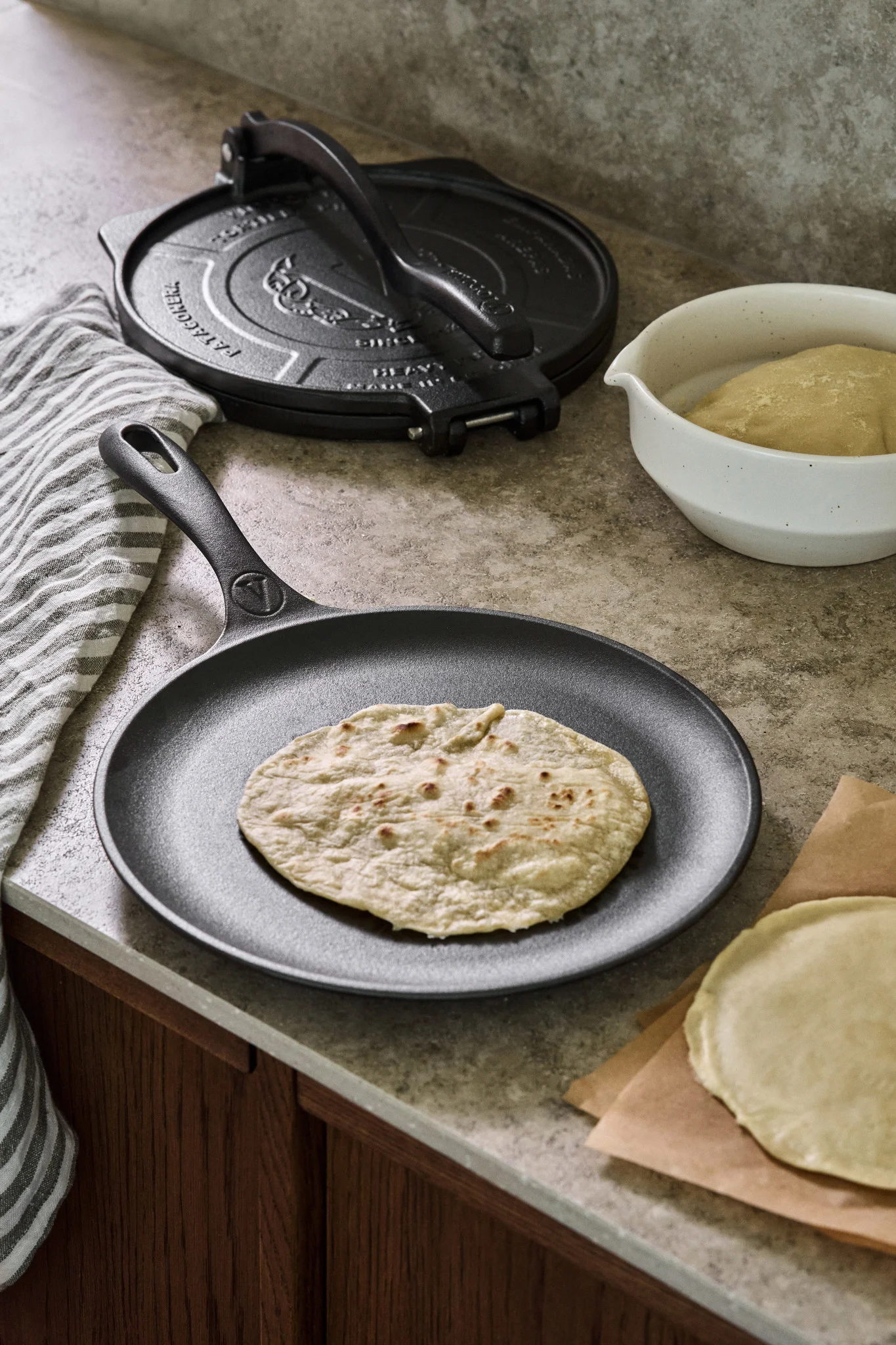 A cast iron skillet on a kitchen side with a fried tortilla, next to it is a cast iron tortilla press and a bowl of dough. 