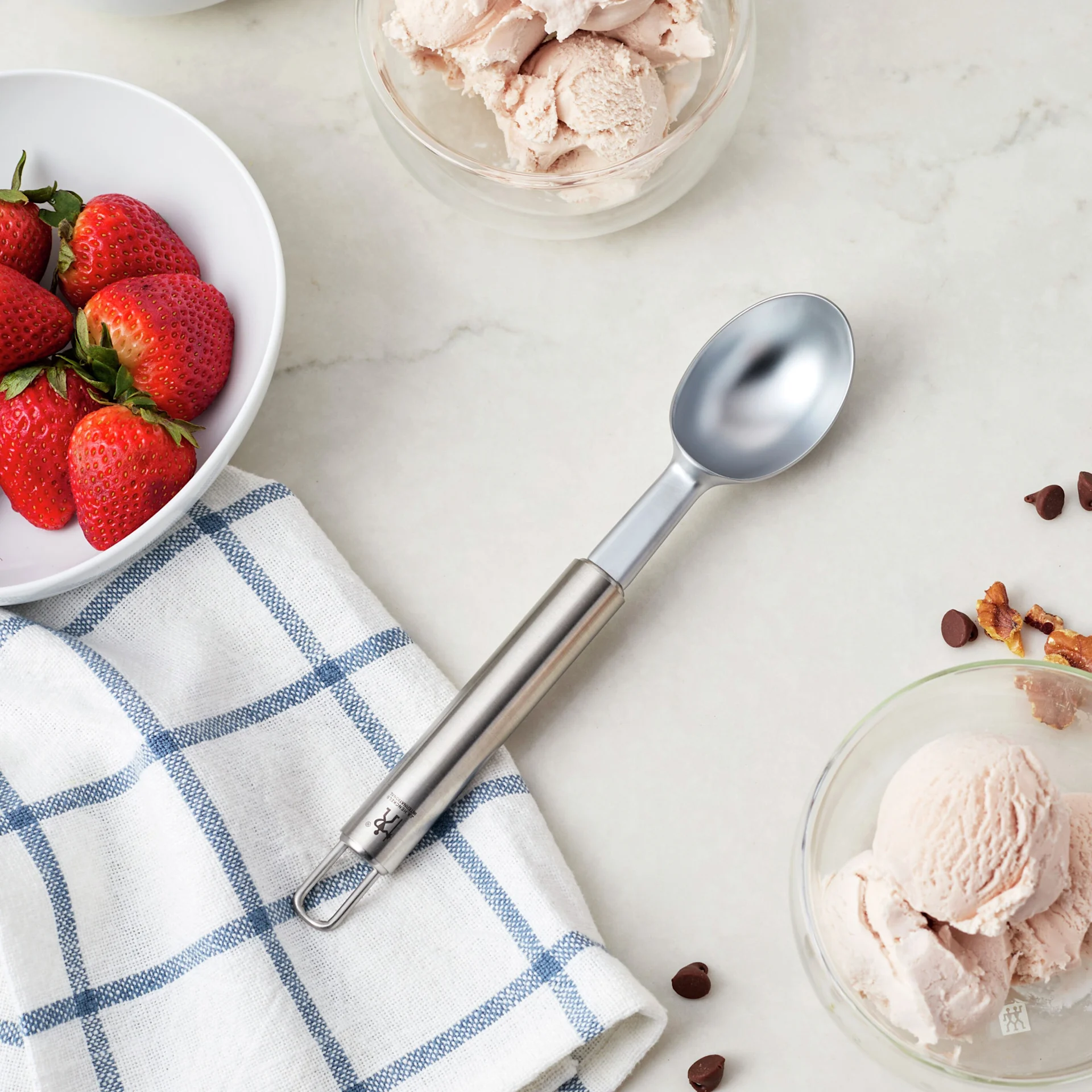 Metal ice cream scoop, fresh strawberries, and two bowls of pink ice cream with toppings on a marble surface.