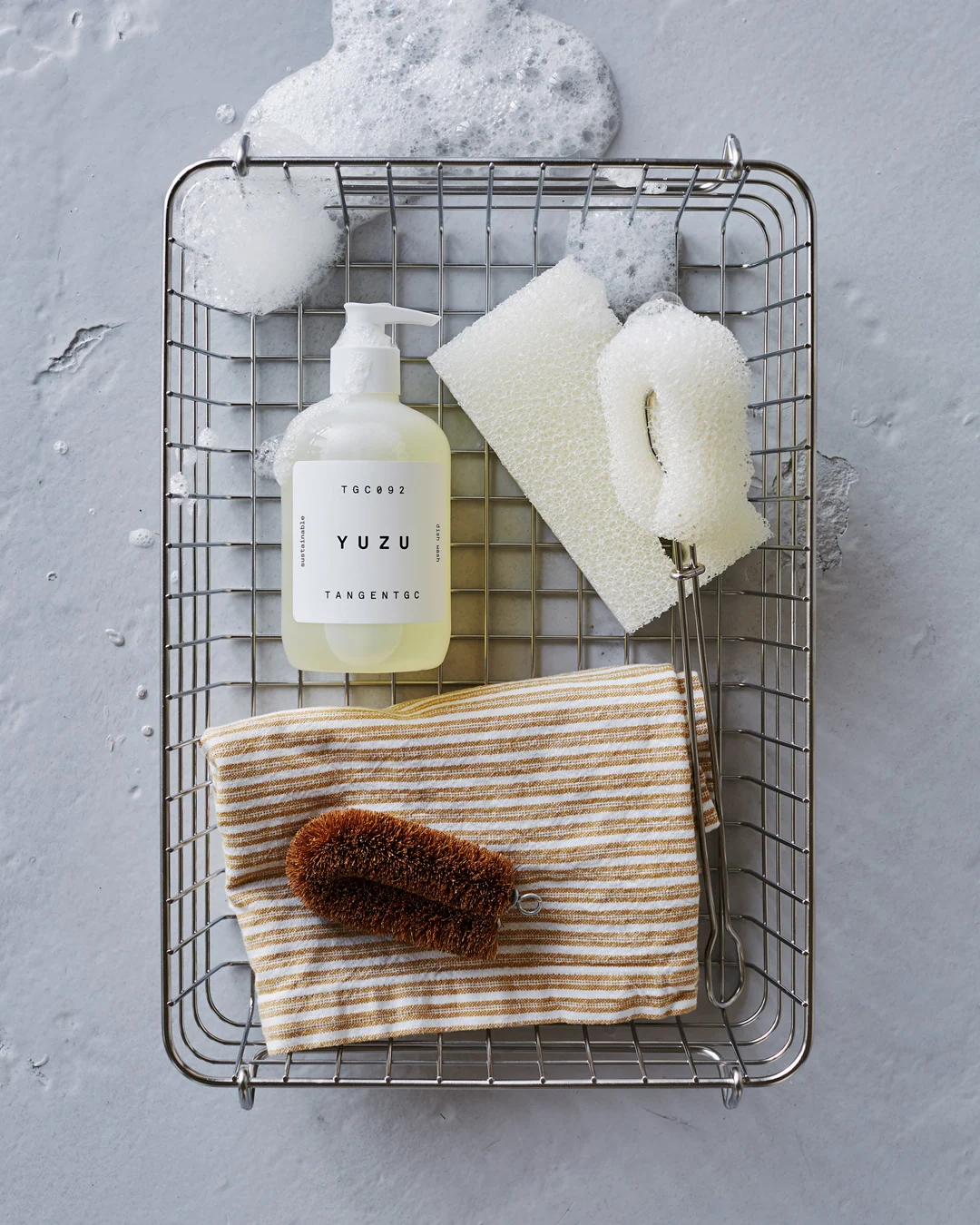 Dish soap, sponges, cloth, and brush in a metal basket with suds on a grey surface.