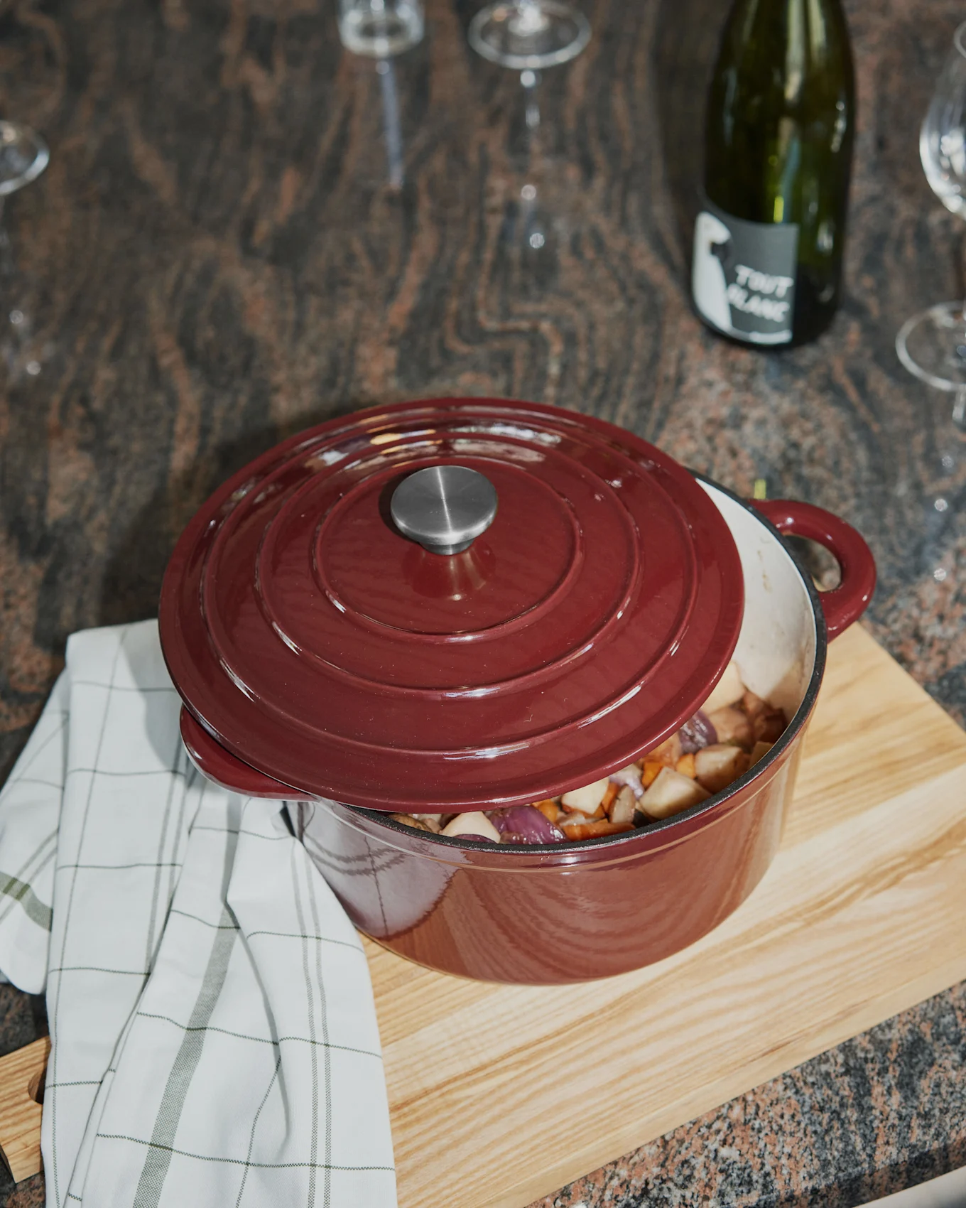 Red cast iron pot with chopped vegetables, lid askew, on a wooden board with a checkered towel.