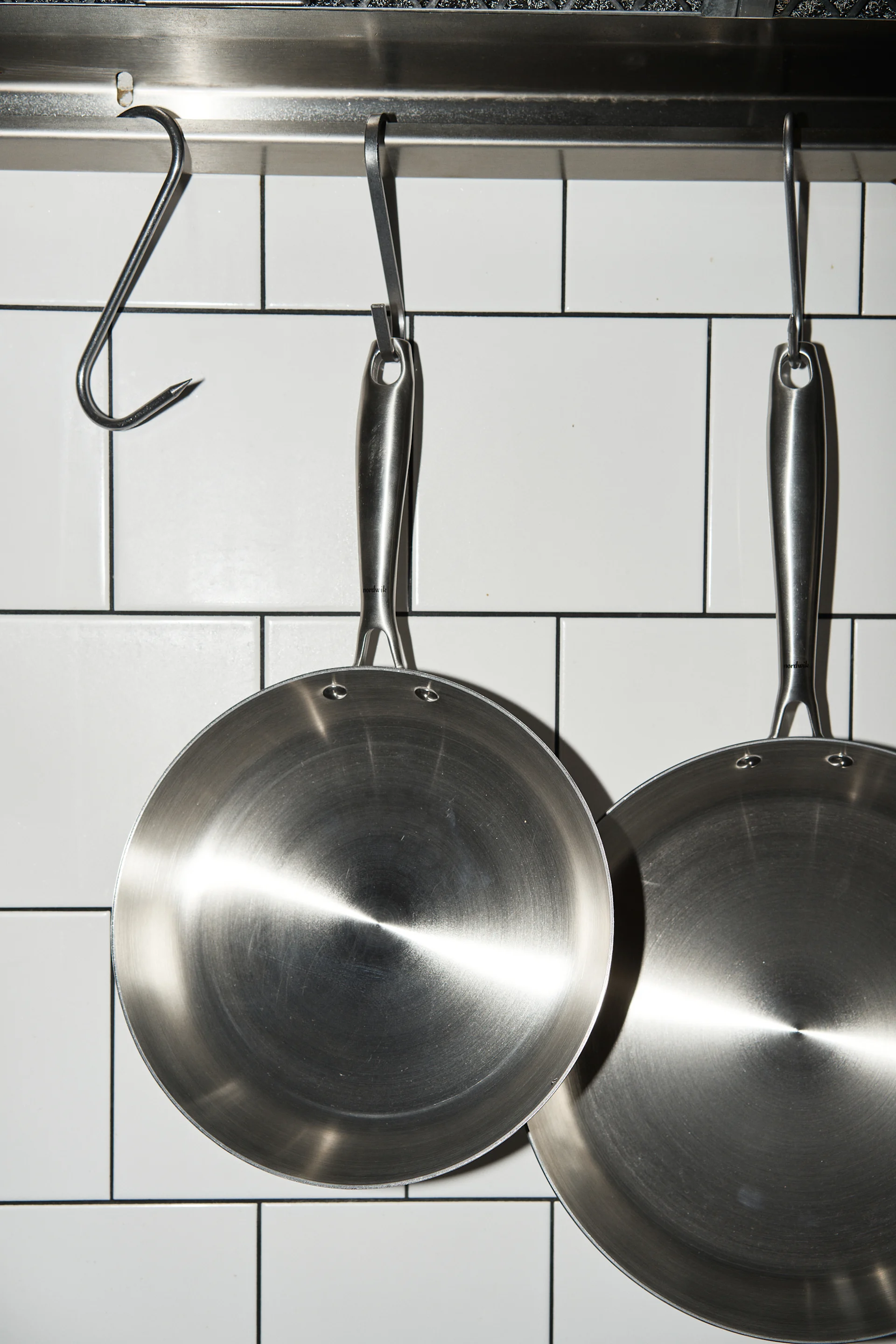 Two stainless steel frying pans hanging on hooks against white subway tiles in a kitchen.