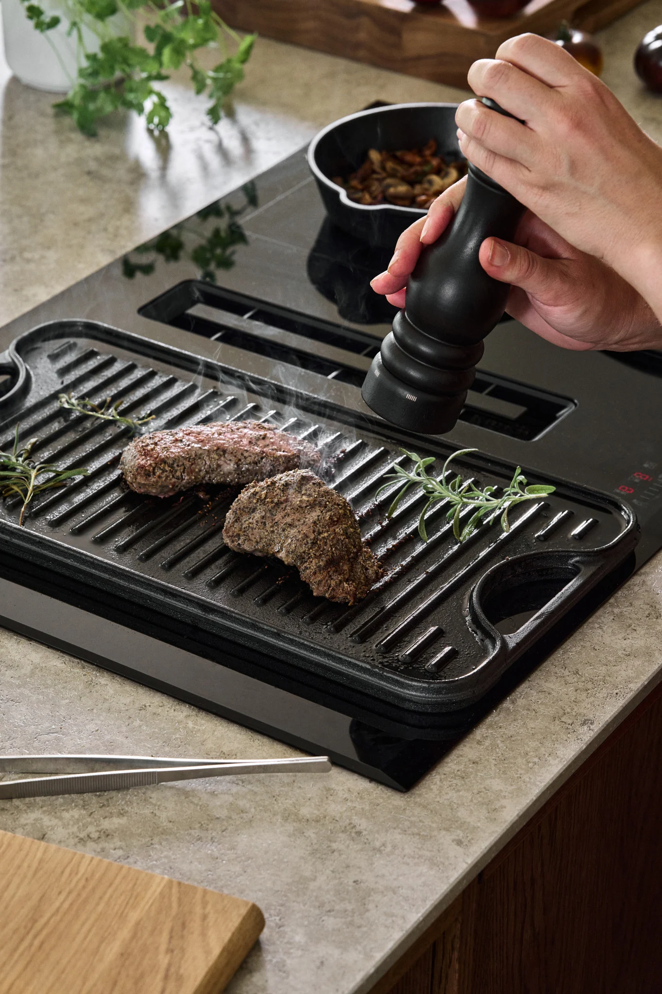 A cast iron cooking plate on an induction hob with a couple of pieces of steak while someone cracks black pepper from a pepper grinder over them.