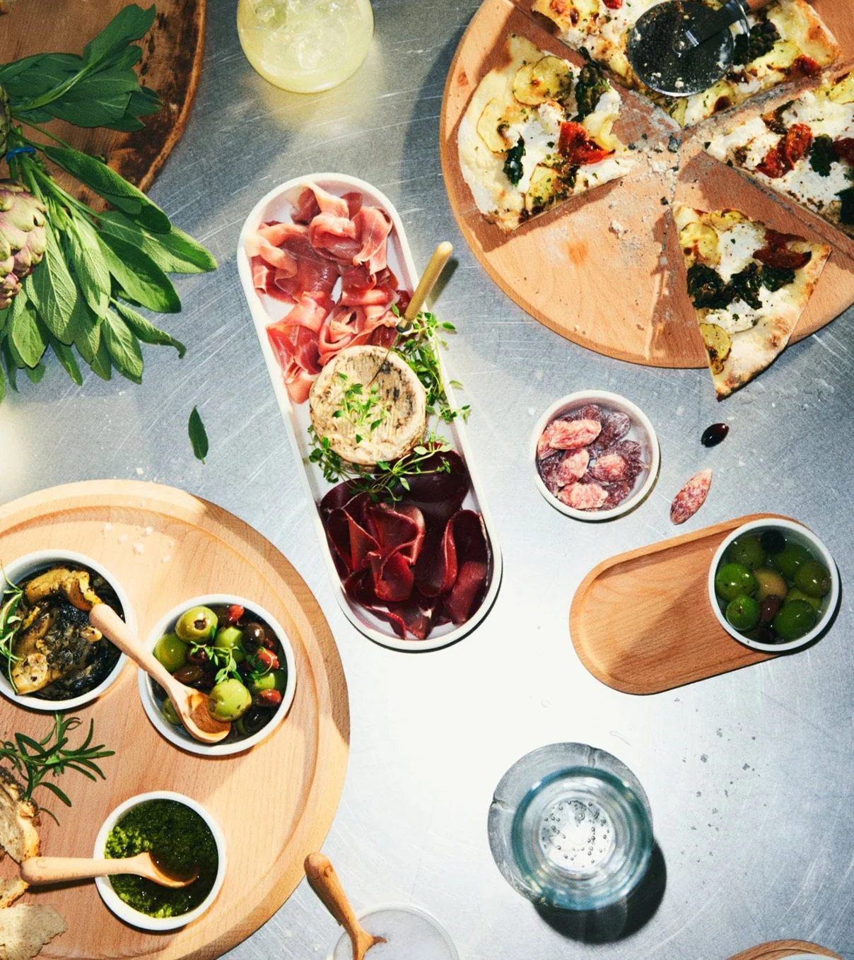 Overhead view of a food spread with a sliced pizza, charcuterie board, olives, pesto, and tapenade on wooden boards.