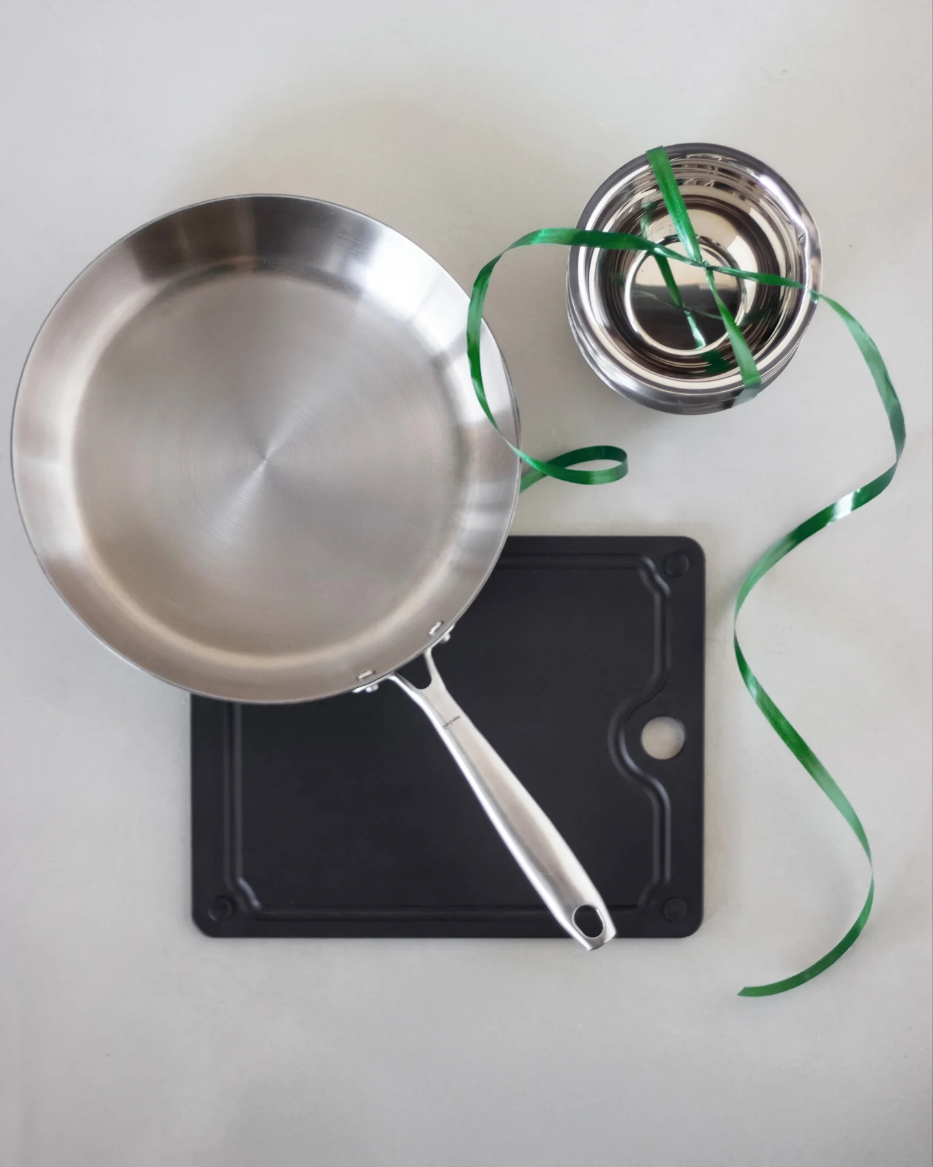 Stainless steel frying pan on a black cutting board, with a stack of steel bowls tied with a green ribbon.