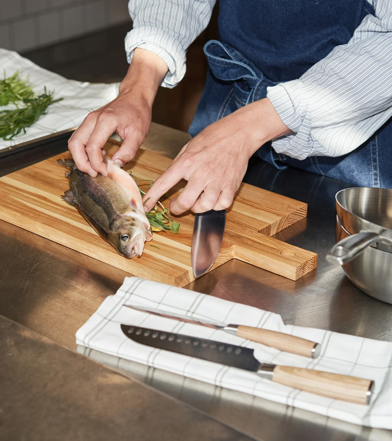 Hands preparing a fish on a wooden cutting board with a knife and fresh herbs. Two knives lie on a towel.