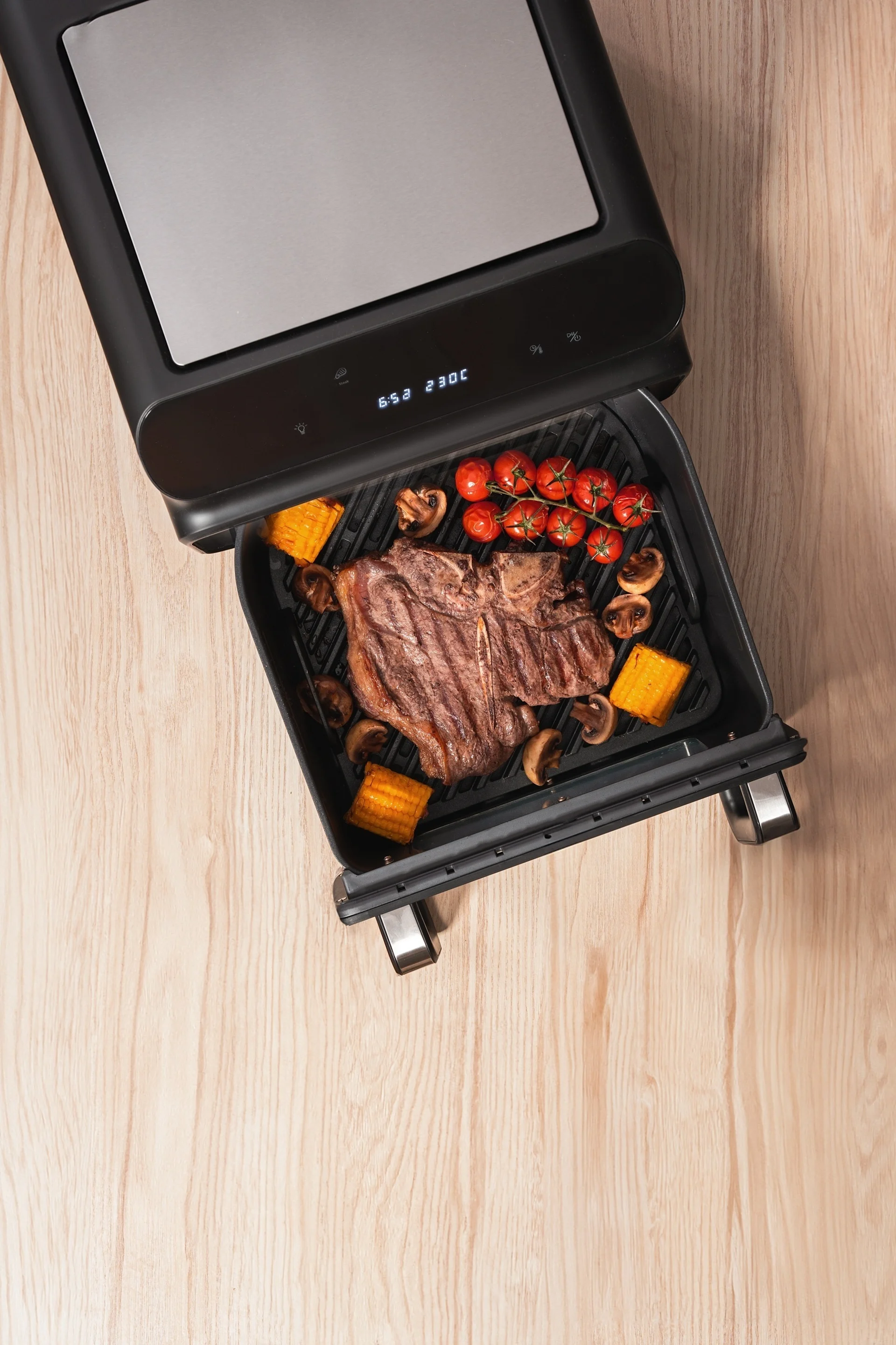 A T-bone steak, cherry tomatoes, mushrooms, and corn on the cob cooking in an open electric grill, displaying 230C.