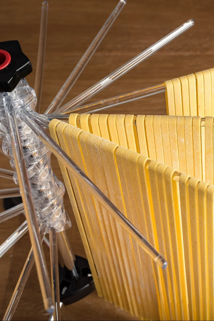 Fresh yellow pasta drying on a clear pasta rack with multiple arms, against a wooden background.
