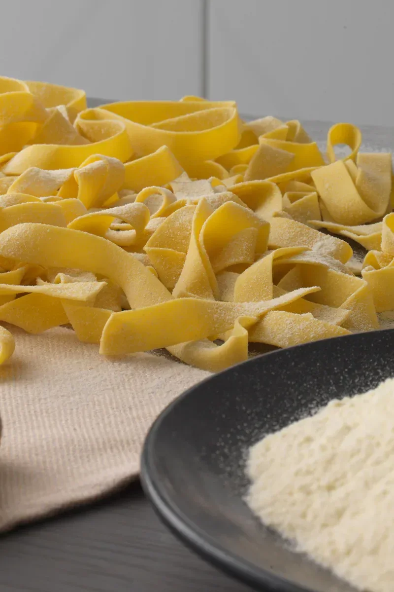 Raw, wide yellow pasta noodles dusted with flour, next to a black plate with more flour.