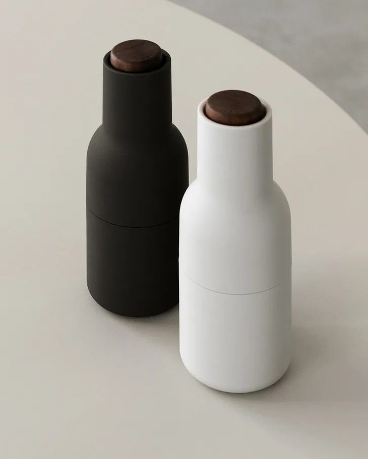 Two elegant bottle-shaped grinders, one black and one white, both with dark wooden tops, on a light surface.