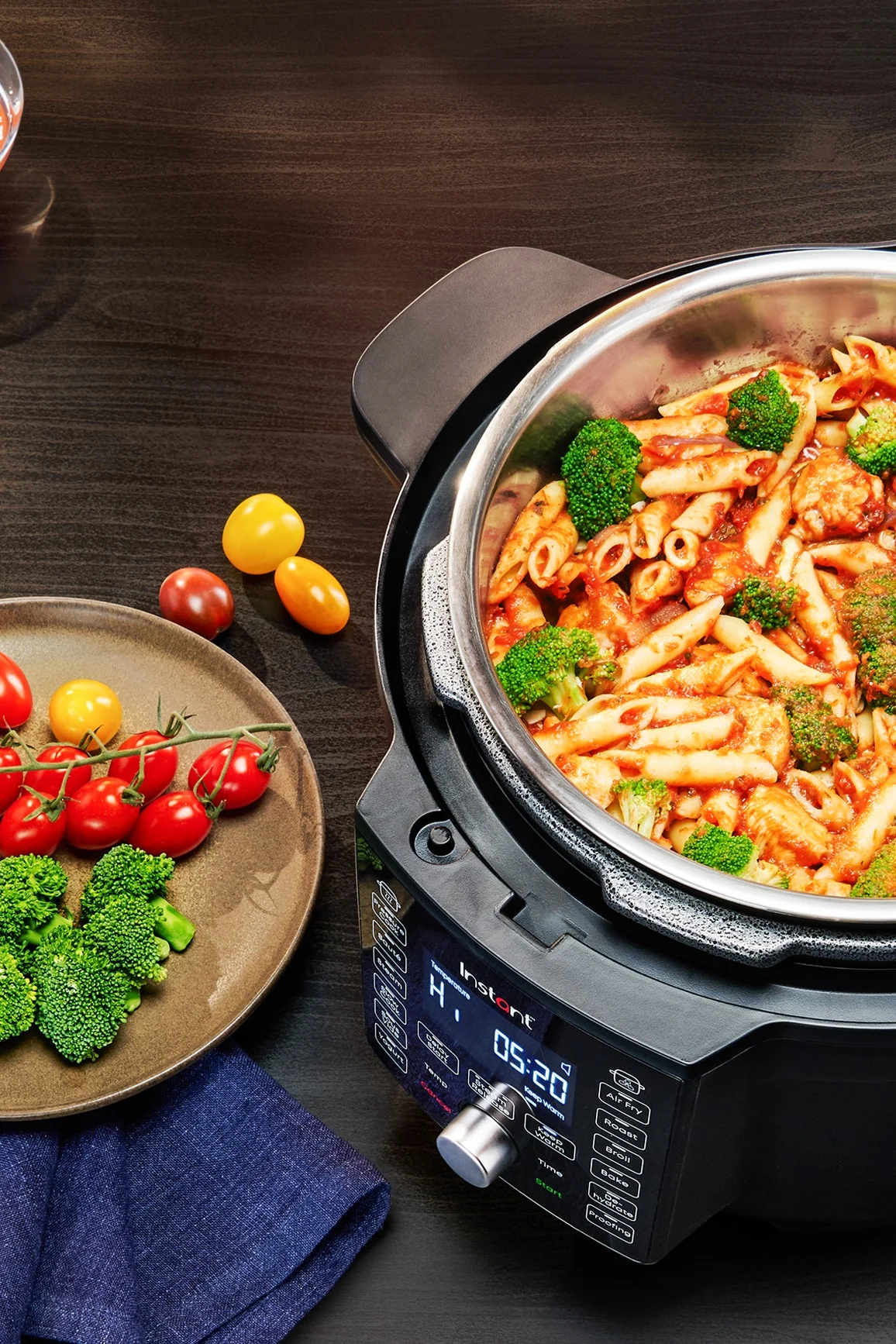 A black multi-cooker filled with penne pasta and broccoli in tomato sauce, alongside a plate of fresh cherry tomatoes and broccoli on a dark wooden table.