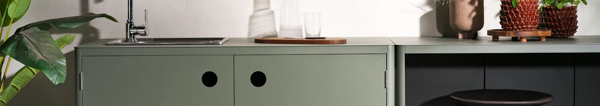 A sage green kitchen area with a sink, cutting board, and potted plants.